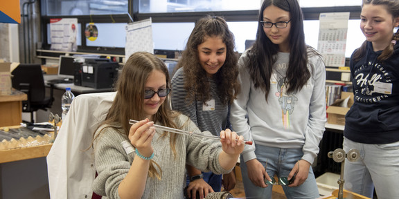 Three girls watch another holding a glass tube over a Bunsen burner.