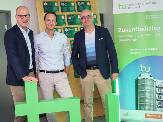 Gruppenfoto: Drei Personen stehen hinter grünen stehenden Buchstaben „TU“. Rechts im Bild steht ein Aufsteller des Zukunftsdialogs.