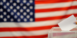 Zu sehen ist die Flagge der USA. Unten rechts ist ein Teil einer durchsichtigen Wahlurne zu sehen, in die gerade ein weißer Zettel eingeworfen wird.