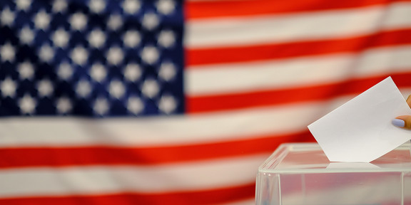 Zu sehen ist die Flagge der USA. Unten rechts ist ein Teil einer durchsichtigen Wahlurne zu sehen, in die gerade ein weißer Zettel eingeworfen wird.
