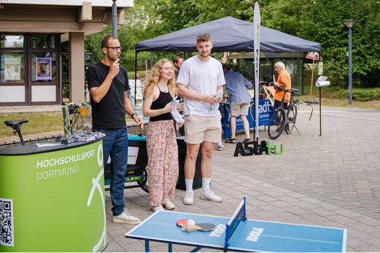 Three people in front of 50 Emil-Figge-Str. at an awards ceremony; in the foreground, a mini table tennis table