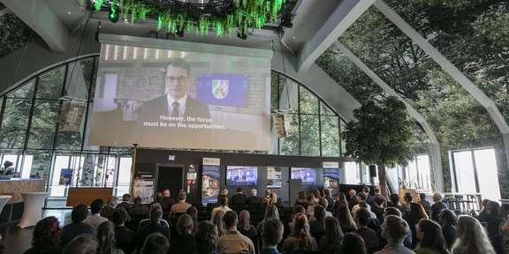 Foto: Ein Veranstaltungssaal im Dortmunder U mit begrünter Decke. Vor dem Publikum wird eine Videobotschaft von Ministerpräsident Wüst abgespielt.