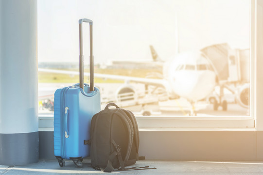 Am Flughafen steht ein blauer Koffer neben einem schwarzen Rucksack und im Hintergrund ist ein Flugzeug auf einem Rollfeld zu sehen.