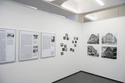 Ecke in einer Ausstellung. Links ein Essay, rechts davon erst ein Cluster kleiner schwarz-weiß Fotos und dann vier schwarz-weiß Architekturfotografien.