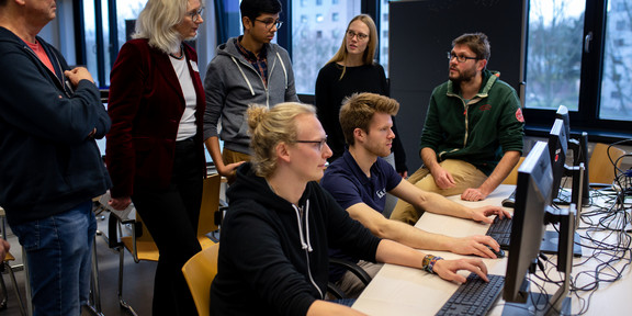 Zwei Personen sitzen an PCs, fünf Leute stehen dahinter und sprechen über etwas, das auf dem Bildschirm zu sehen ist