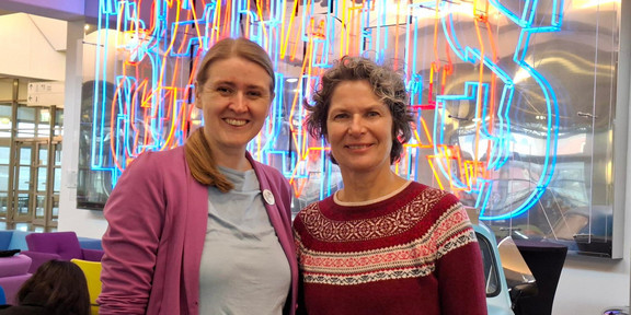 Foto: Prof. Doris Reiter und Dr. Christiane Wüllner stehen in einem Raum vor bunter Leuchtschrift.