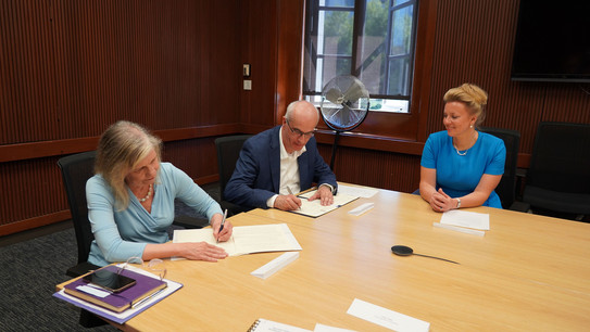 An einem Konferenztisch unterschreiben eine Frau mit grauen Haaren und Rektor Manfred Bayer im dunkelblauen Jacket eine Urkunde. Rechts von Herrn Bayer sitzt Wissenschaftsministerin Ina Brandes im blauen Kleid und lächelt ihnen zu.