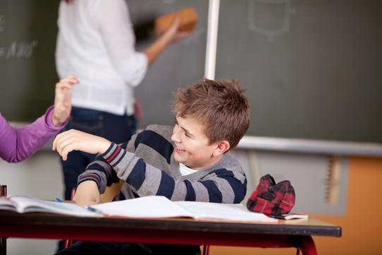 Ein junger Schüler sitzt an seinem Tisch im Klassenzimmer und albert mit seinem Sitznachbarn herum.