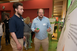 Two men are standing in front of a poster, talking to each other. In the background, other people are standing in front of different posters.
