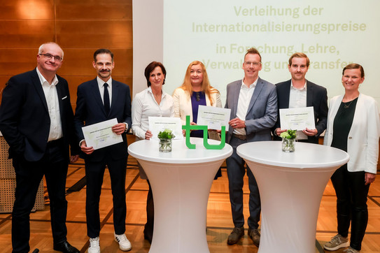 Seven people stand around two bar tables and pose for a photo. The five people in the middle are holding prize certificates in their hands.