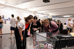 Drei Personen schauen in einen Einkaufswagen. Dahinter schauen sich Menschen die Ausstellung an.
