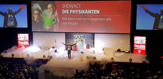 Eine große Bühne, die mit Nebel bedeckt ist. Ein Mann steht in der Mitte und hält die Hände nach oben. Im Hintergrund ist ein Banner mit der Aufschrift "Showact - Die Physikanten" zu sehen.