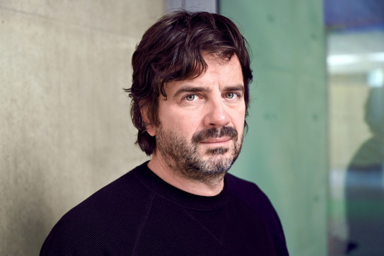 Porträtfoto: Ein Foto von Prof. Nikola Ukić in einem schwarzen Pullover vor einem verschwommenen grünen und braunen Hintergrund.