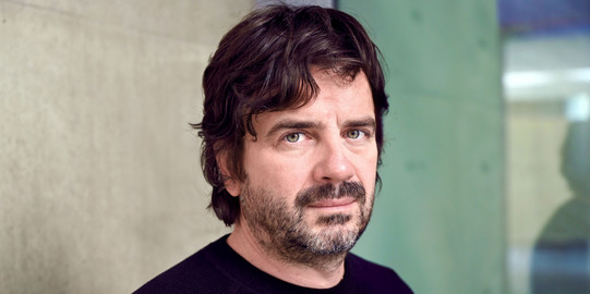 Porträtfoto: Ein Foto von Prof. Nikola Ukić in einem schwarzen Pullover vor einem verschwommenen grünen und braunen Hintergrund.