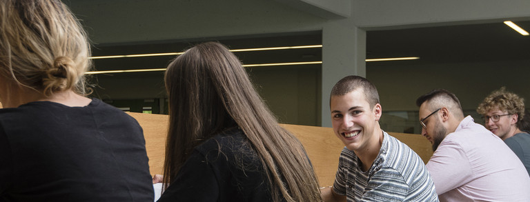 Studierende_Rueckenansicht.jpg Fünf Studierende sitzen nebeneinander an einem langen Tisch und arbeiten. Der mittlere Student schaut in die Kamera, die anderen haben dem Betrachter den Rücken zugewandt