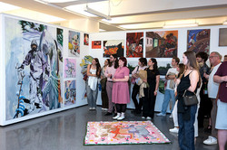 Mehrere Besucher*innen der Galerie, die vor einer Wand mit vielen Kunstwerken stehen. Vor den Besuchenden liegt ein weiteres Kunstwerk auf dem Boden.