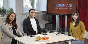 Eine Frau sitzt am Tisch und interviewt einen Mann und eine Frau. Auf einem Bildschirm im Hintergrund steht der Titel der neuen Sendung.