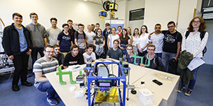 Gruppenfoto der Schülerinnen und Schüler, auf Tisch im Vordergrund stehen ein Versuchsaufbau und das grüne TU-Logo