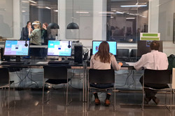 Das Bild zeigt eine Tischreihe mit Bildschirmcomputern, die an einer Glaswand stehen. Zwei Personen sitzen in schwarzen Stühlen vor diesen Bildschirmen mit dem Rücken zur Kamera. Hinter der Glaswand befindet sich ein weiterer Raum.
