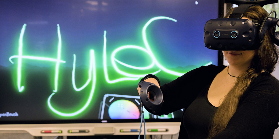 Eine Frau hat eine VR-Brille auf und hält in jeder Hand einen kleines Steuerinstrument für die VR-Umgebung. Im Hintergrund ist ein Bildschirm zu sehen, auf dem der Schriftzug Hylec eingeblendet ist.. 