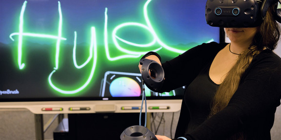 Eine Frau hat eine VR-Brille auf und hält in jeder Hand einen kleines Steuerinstrument für die VR-Umgebung. Im Hintergrund ist ein Bildschirm zu sehen, auf dem der Schriftzug Hylec eingeblendet ist..