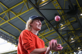 Ein Mann mit Kappe jongliert drei Bälle auf einer überdachten Tribüne in einem Fußballstadion.