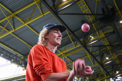 A man in a cap juggles three balls on a covered stand in a soccer stadium.