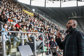 Ein Mann mit Mikrofon in der Hand steht vor einer mit Erstsemesterstudierenden gefüllt Tribüne im Stadion und spricht/singt ins Mikrofon.