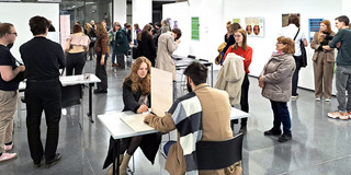 Das Bild zeigt einen Ausstellungsraum mit Besucher:innen. Im Zentrum des Bildes sitzen sich zwei Menschen an einem Tisch gegenüber. Auf dem Tisch selbst steht eine Trennwand. Die Personen haben ihre eigenen Unterarme jeweils auf dem Tisch auf der einen und der anderen Trennseite aufgelegt. Im Hintergrund sind weitere Tische und Besucher:innen zu sehen sowie eine weiße Wand mit Leinwänden.