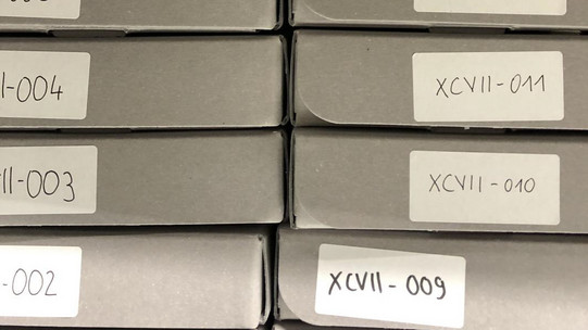 Nahaufnahme von zwölf grauen Archivboxen mit nummerierten Aufklebern, die übereinander gestapelt sind.