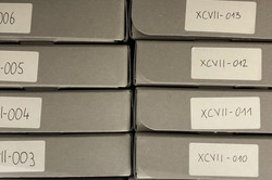Nahaufnahme von zwölf grauen Archivboxen mit nummerierten Aufklebern, die übereinander gestapelt sind.