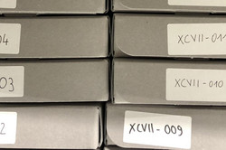 Close-up of twelve gray archive boxes with numbered stickers stacked on top of each other.