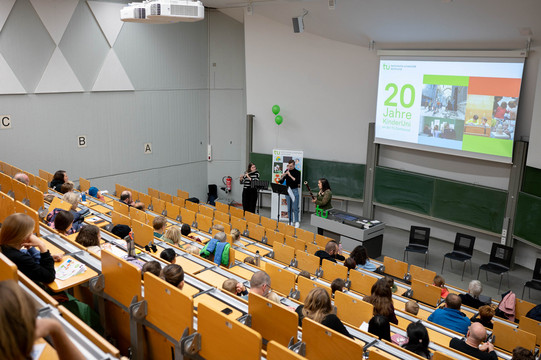 In einem Hörsaal spielen eine Frau mit Querflöte, ein Mann mit Oboe und eine Frau mit Fagott Musik. Im Publikum sitzen Kinder und ihre Familien. Auf einer Präsentation steht: „20 Jahre KinderUni an der TU Dortmund“.