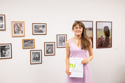 Im Zentrum des Bildes befindet sich Marie Schütte, die mit ihrem Preis-Zertifikat vor einer weißen Wand mit fotografischen Kunstwerken steht und in die Kamera lächelt.