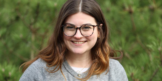 Mitarbeiterin Emily Weidle steht im Freien vor einer grünen Hecke