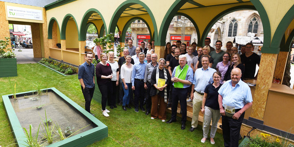 Group photo in the "stadt paradies sanktreinoldi: The people stand partly in the green-yellow painted arcade, partly on the planted lawn