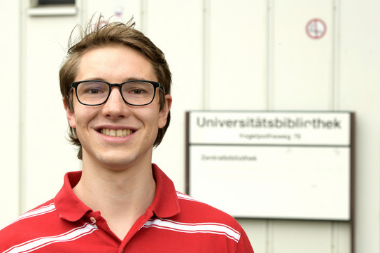 Fabian Dillenhöfer Ein Mann steht vor der Universitätsbibliothek und lächelt in die Kamera