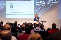 Foto: Prof. Dirk Schadendorf, Gründungsdirektor des Research Center One Health Ruhr, steht vor dem Publikum an einem Redepult. Hinter ihm ist eine Präsentation zu sehen.