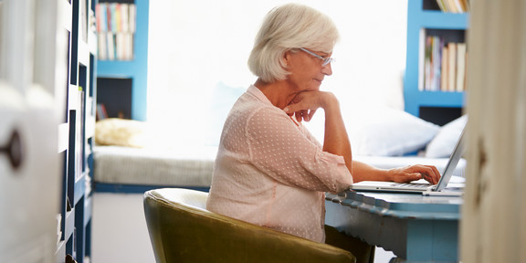 Weiterbildendes Studium für Seniorinnen und Senioren Eine ältere Frau mit weißen Haaren sitzt an einem Tisch und arbeitet an ihrem Laptop. Im Hintergrund stehen Bücherregale und eine Couch.