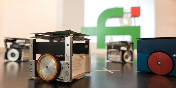 Kleine würfelförmige Roboter und das Logo der Fakultät für Informatik Im Vordergrund kleine würfelförmige Roboter, im Hintergrund das Logo der Fakultät für Informatik