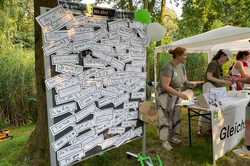 Eine große Magnettafel auf der viele Sprechblasen angeheftet sind. Auf den Sprechblasen steht "Ich bin TU" in verschiedenen Sprachen. Rechts sieht man mehrere Frauen vom Gleichstellungsbüro unter einem Pavillon.