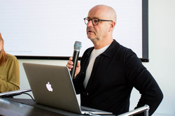 Das Bild zeigt eine männlich gelesene Person im schwarzen Jackett hinter einem silbernen Apple-Laptop. In seiner rechten Hand hält er ein Mikrophon, in das er spricht. Außerdem trägt er eine schwarze Brille und eine Glatze.