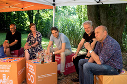 Photo: Several people sit under a pavilion and take part in a discussion at an outdoor event.