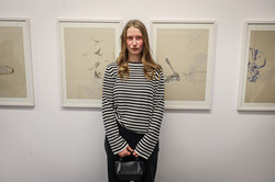 Foto: Eine junge Frau mit blonden Haaren steht vor einer Bildergalerie und schaut in die Kamera. In den Händen hält sie eine Tasche.
