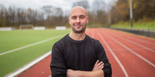 Portrait of Prof. Dennis Dreiskämper: A man in a black sweater stands with his arms folded on a running track on campus.