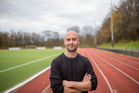 Portrait of Prof. Dennis Dreiskämper: A man in a black sweater stands with his arms folded on a running track on campus.
