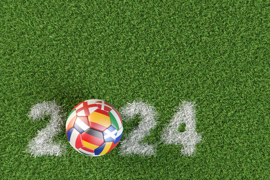 Foto: Fußball mit den Flaggen mehrerer Länder, die sich für die Europameisterschaft qualifiziert haben. Die Jahreszahl 2024 als Kreidemarkierung auf dem Rasen.