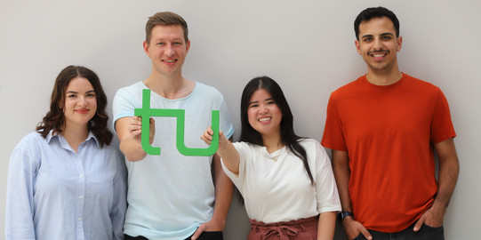 Vier Studierende stehen in einer Reihe. In der MItte halten zwei Studierende ein TU Dortmund-Logo aus Holz.