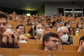 Mehrere Personen sitzen in einem Hörsaal. Einige Personen tragen Masken.