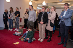 Das Bild zeigt eine Traube von Menschen auf einem roten Teppich stehend und haben ihre Aufmerksamkeit in die gleiche Richtung gerichtet. Drei Kinder sitzen in erster Reihe auf dem Teppich.